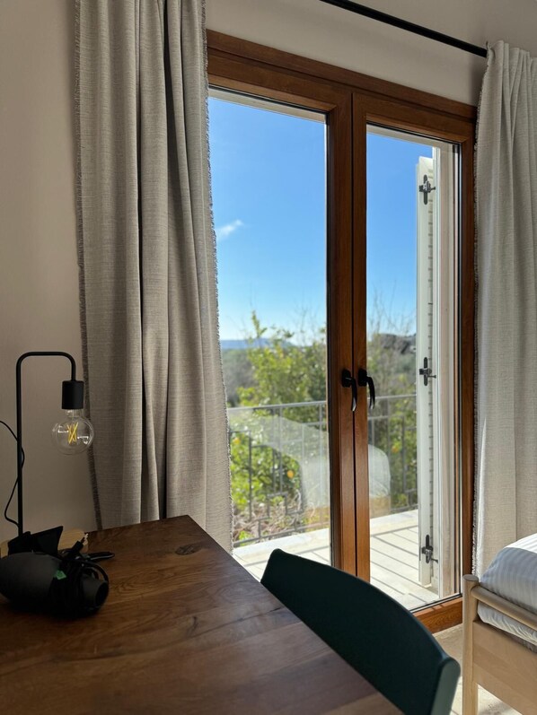 A room with a wooden table, a green chair, a lamp, and a sliding glass door leading to an outdoor area with a view of greenery and a clear sky.