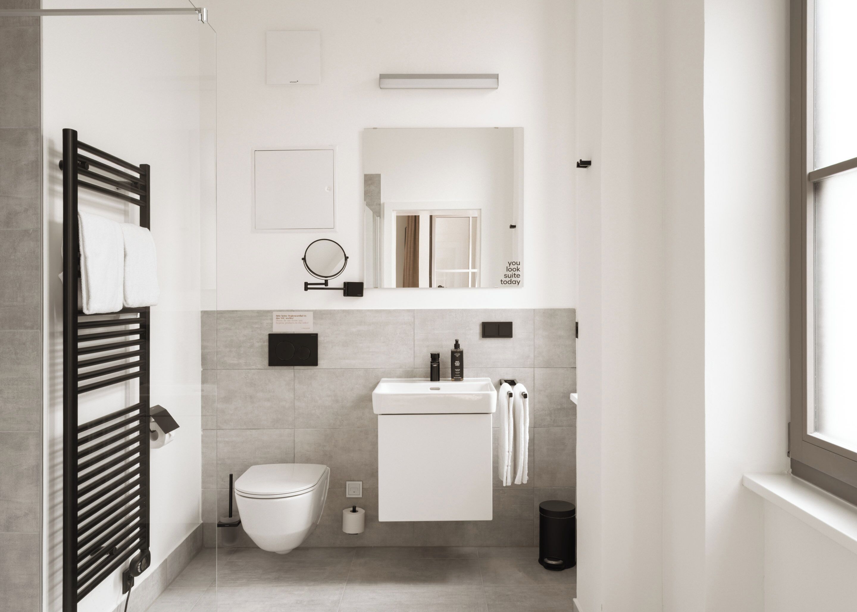 A modern bathroom with a toilet, sink, mirror, towel rack, and a black trash bin.