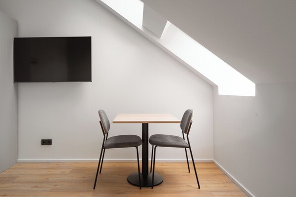 Una habitación moderna con piso de madera, una mesa redonda, dos sillas grises, un televisor de pantalla plana montado en la pared y un tragaluz.