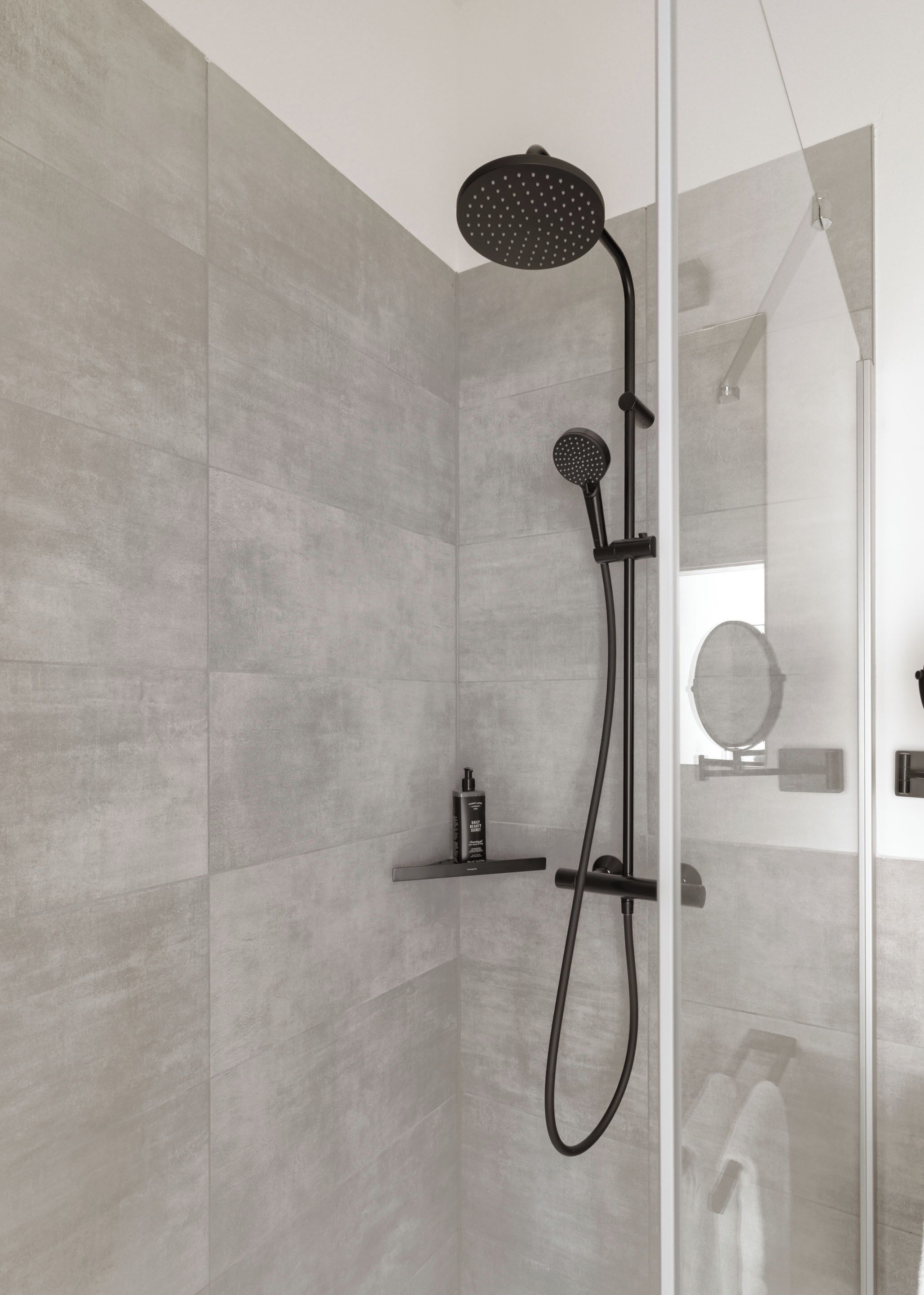 A modern shower with a handheld showerhead and a fixed wall-mounted head, a glass door, and a small shelf with toiletries.