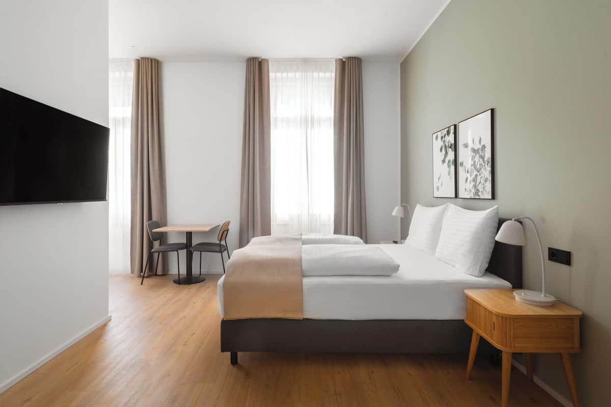 A modern hotel room with a bed, bedside table, lamp, and a TV mounted on the wall.
