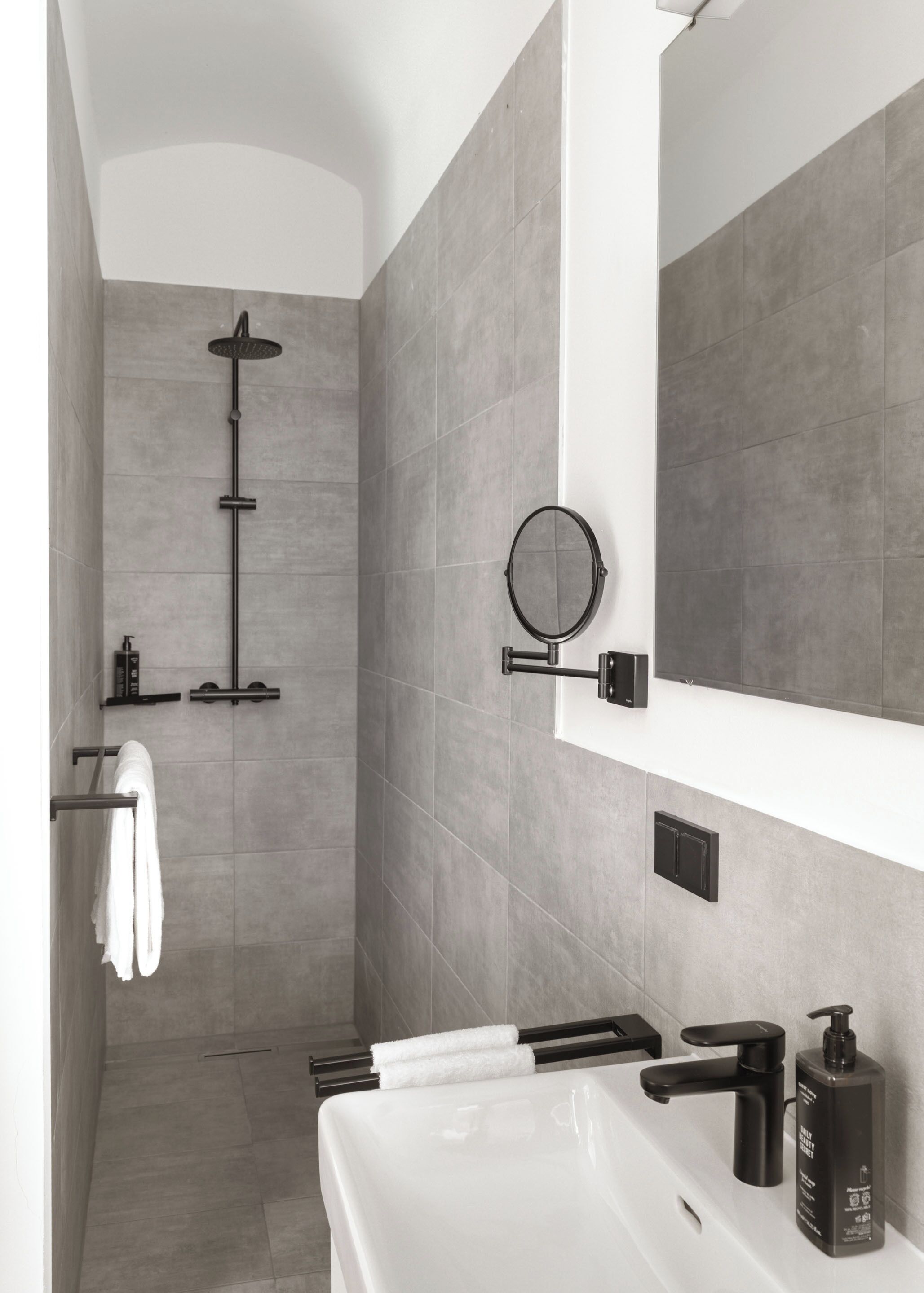 Un baño moderno con ducha a ras de suelo, un espejo grande, un lavabo blanco y un dispensador de jabón negro.