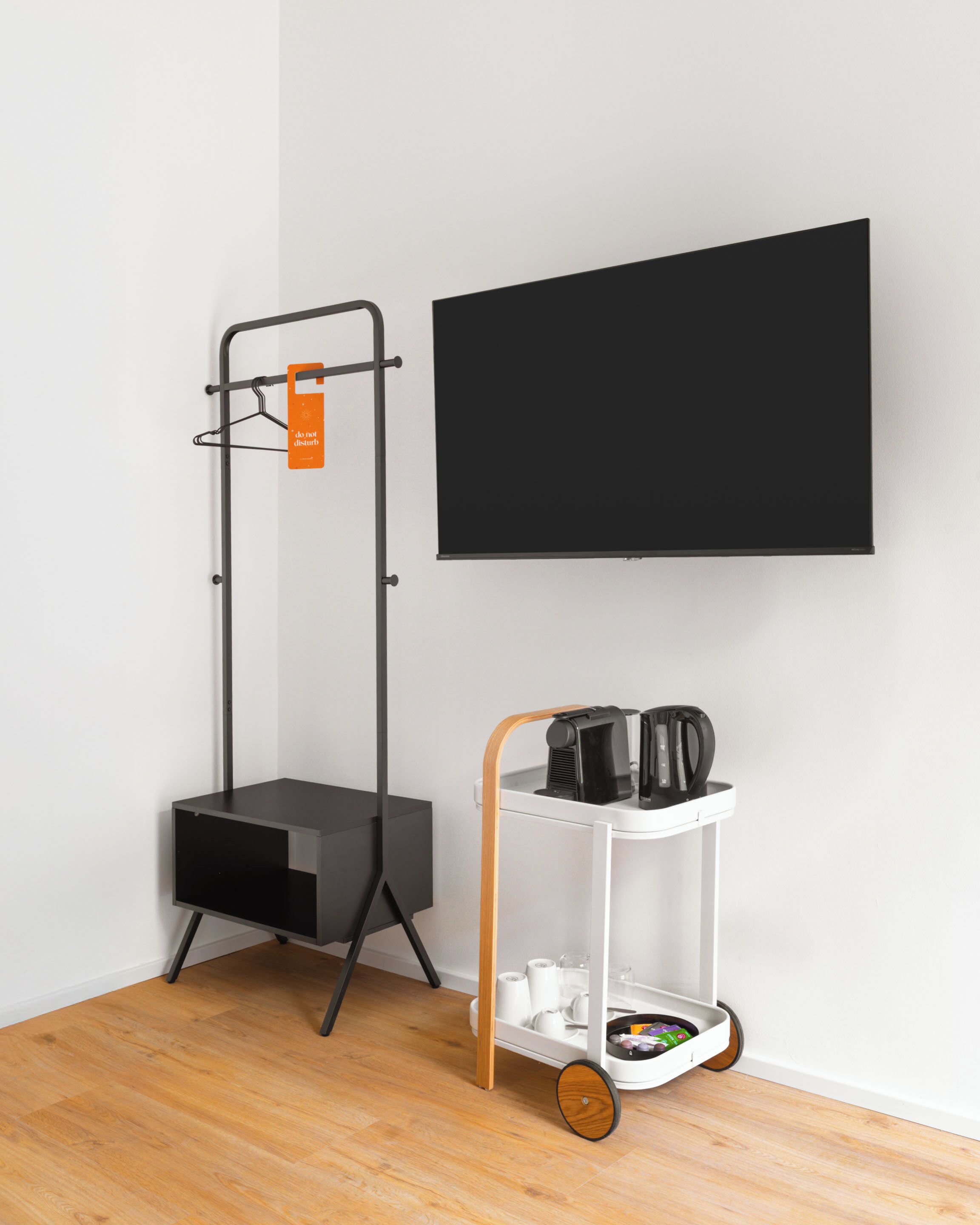 Ein Zimmer mit einem an der Wand befestigten schwarzen Fernseher, einem grauen Stauraum und einem weißen Wagen mit Wasserkocher und Tassen.