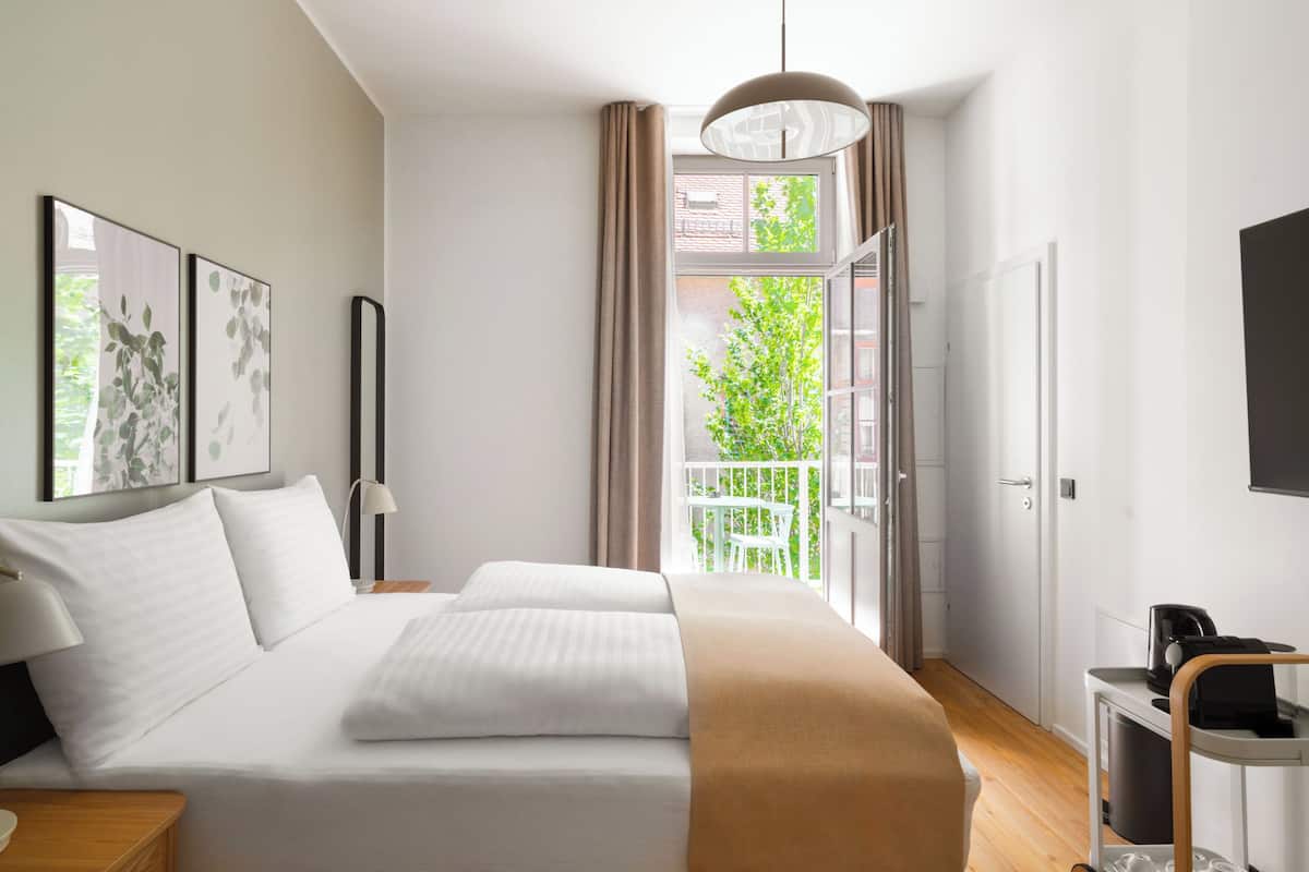 A modern hotel room with a large bed, a bedside table, a lamp, a view of greenery outside, and a balcony.