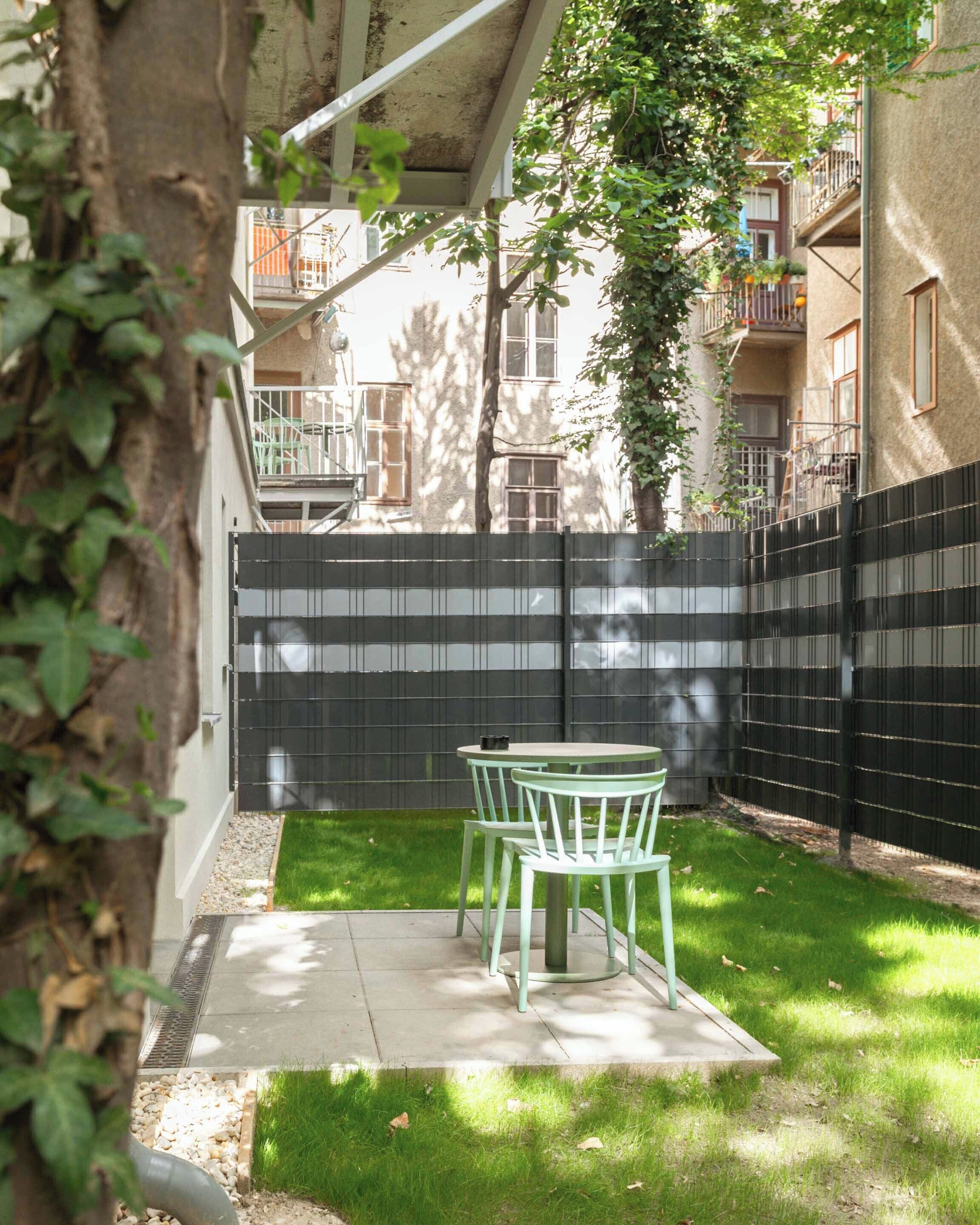 A small outdoor patio with a table and chairs, surrounded by a black fence and a grassy area.
