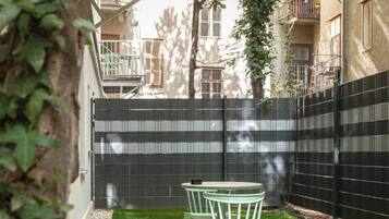 A small outdoor patio with a table and chairs, surrounded by a black fence and a grassy area.
