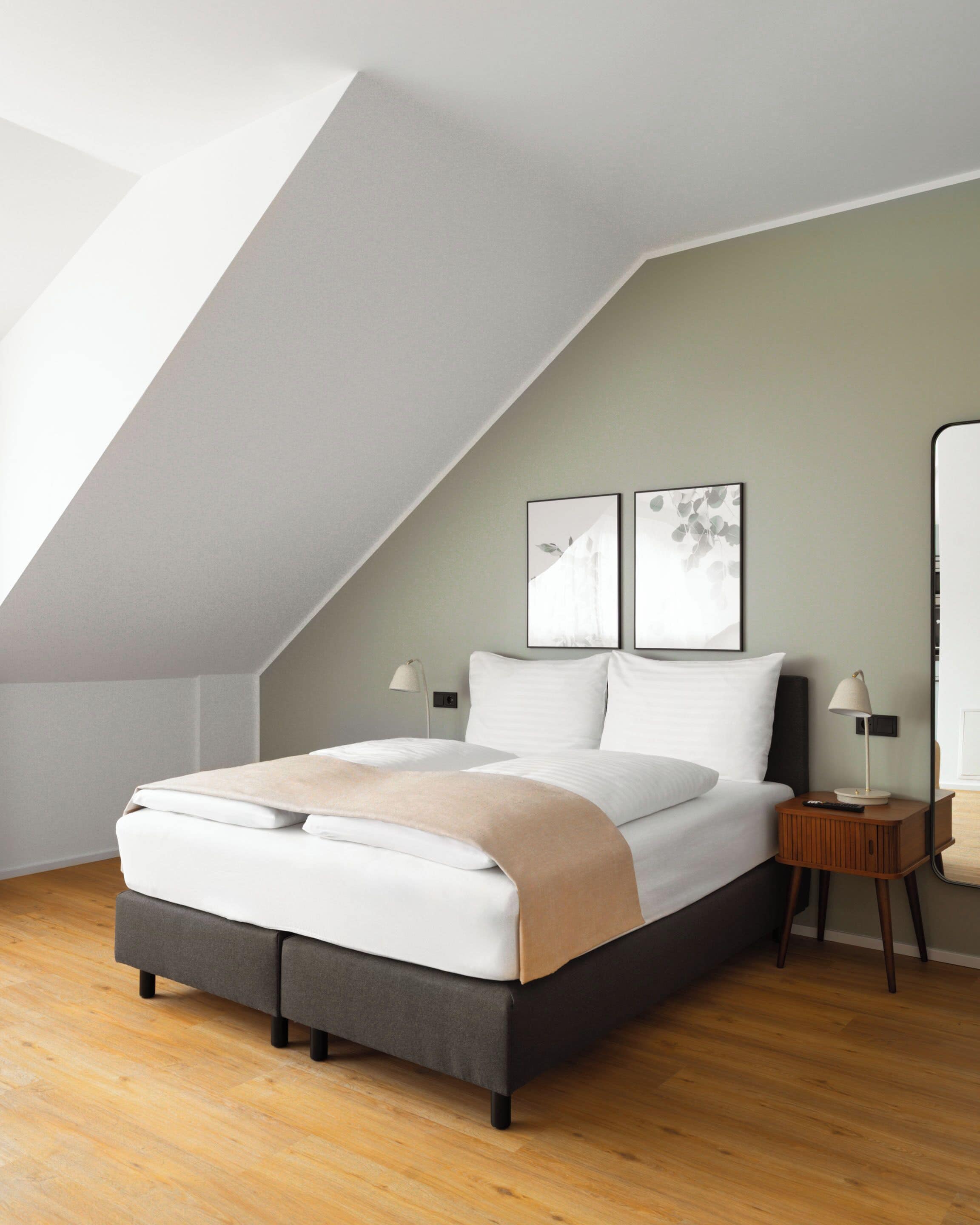 Un dormitorio moderno con techo inclinado, una cama con manta color beige, dos mesitas de noche con lámparas, y dos cuadros enmarcados en la pared.