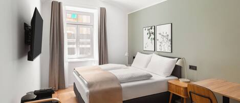 A modern hotel room with a bed, a desk, a chair, a TV mounted on the wall, and artwork on the wall.