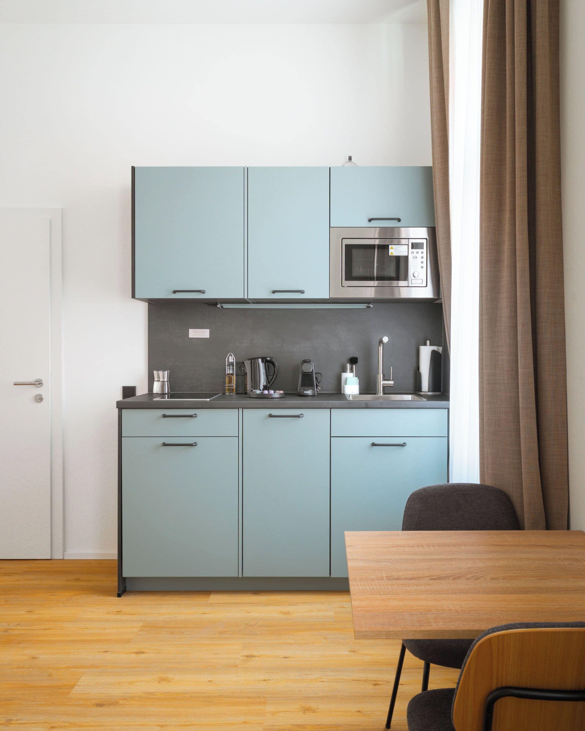 A compact kitchen with light blue cabinets, a microwave, and a coffee maker. There is a wooden table and a chair in the foreground.