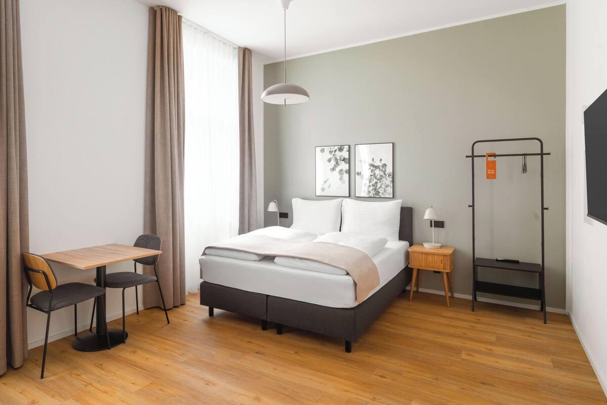 A modern hotel room with a bed, a desk, a chair, a wardrobe, and a hanging light fixture.