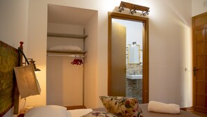 A bedroom with a bed, floral pillows, a hanging rack with clothes, a wooden door, and a mirror reflecting a bathroom. - Villa I 2 Pini (San Vito Lo Capo)