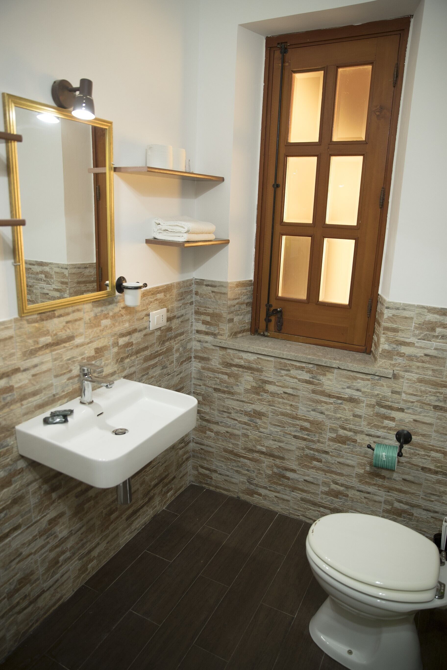 Une salle de bain avec un lavabo blanc, des toilettes, un miroir avec un cadre doré et une porte en bois dotée de panneaux de verre.
