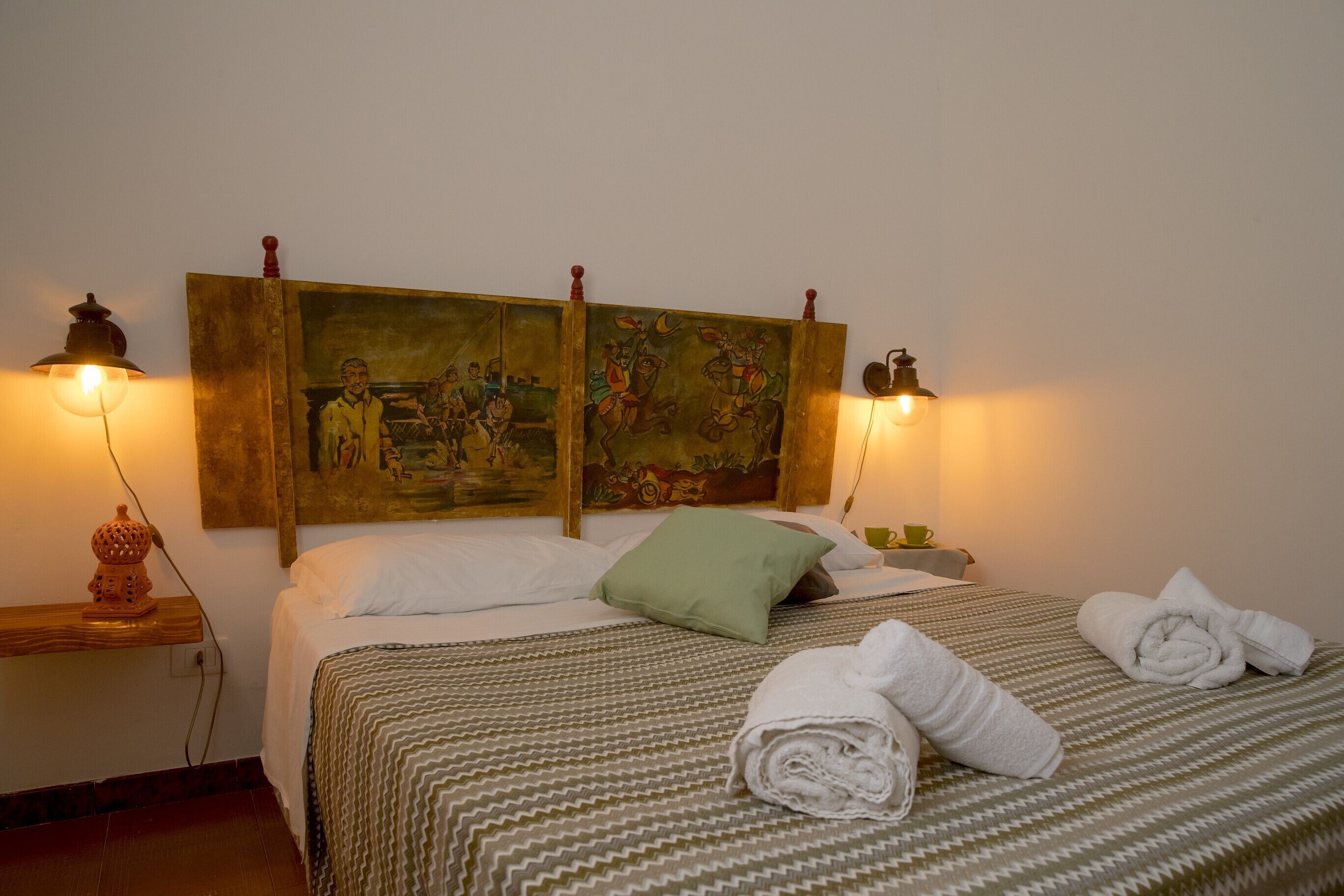 A neatly made bed with a patterned bedspread, a green pillow, and rolled towels on a wooden headboard with paintings.