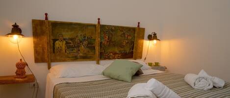 A neatly made bed with a patterned bedspread, a green pillow, and rolled towels on a wooden headboard with paintings.