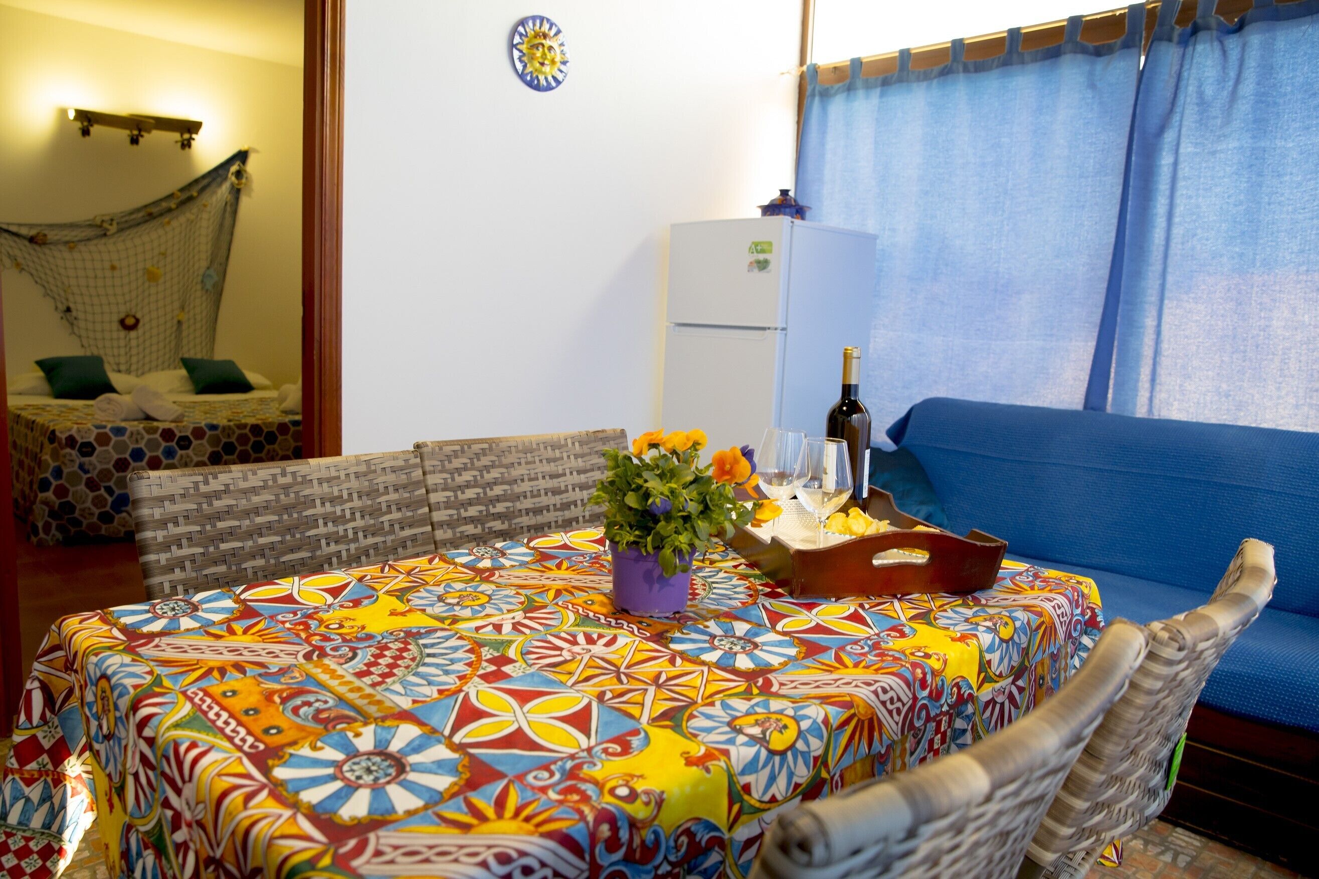 Una habitación con una mesa redonda cubierta por un mantel colorido, un sofá de mimbre, una hamaca y un refrigerador.