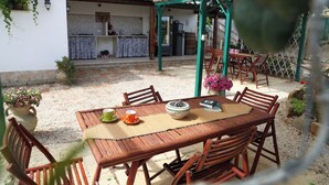 An outdoor patio with a wooden table and chairs, a thatched roof, and a kitchen area in the background. - Villa I 2 Pini (San Vito Lo Capo)