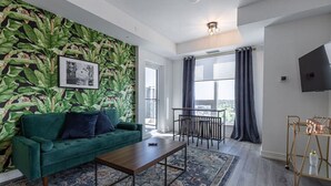 A modern living room with a green sofa, a coffee table, and a patterned rug. There is a large window with curtains, a TV mounted on the wall, and a plant wall. - Executive 2BD+Wifi+Parking+EV (Kitchener)
