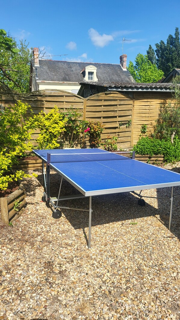 A ping pong table in a garden area with a wooden fence, gravel ground, and a house in the background. - Gîte "le charme (Cinais)