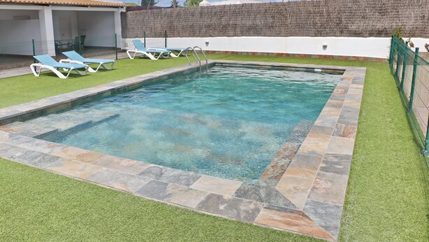 A rectangular swimming pool with a stone border, surrounded by artificial grass and equipped with blue lounge chairs.