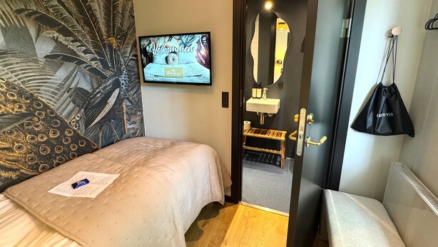 A hotel room with a bed, a television, a mirror, a small table, and a bag hanging on the wall. - Hotel Arnkilhus (Sønderborg)