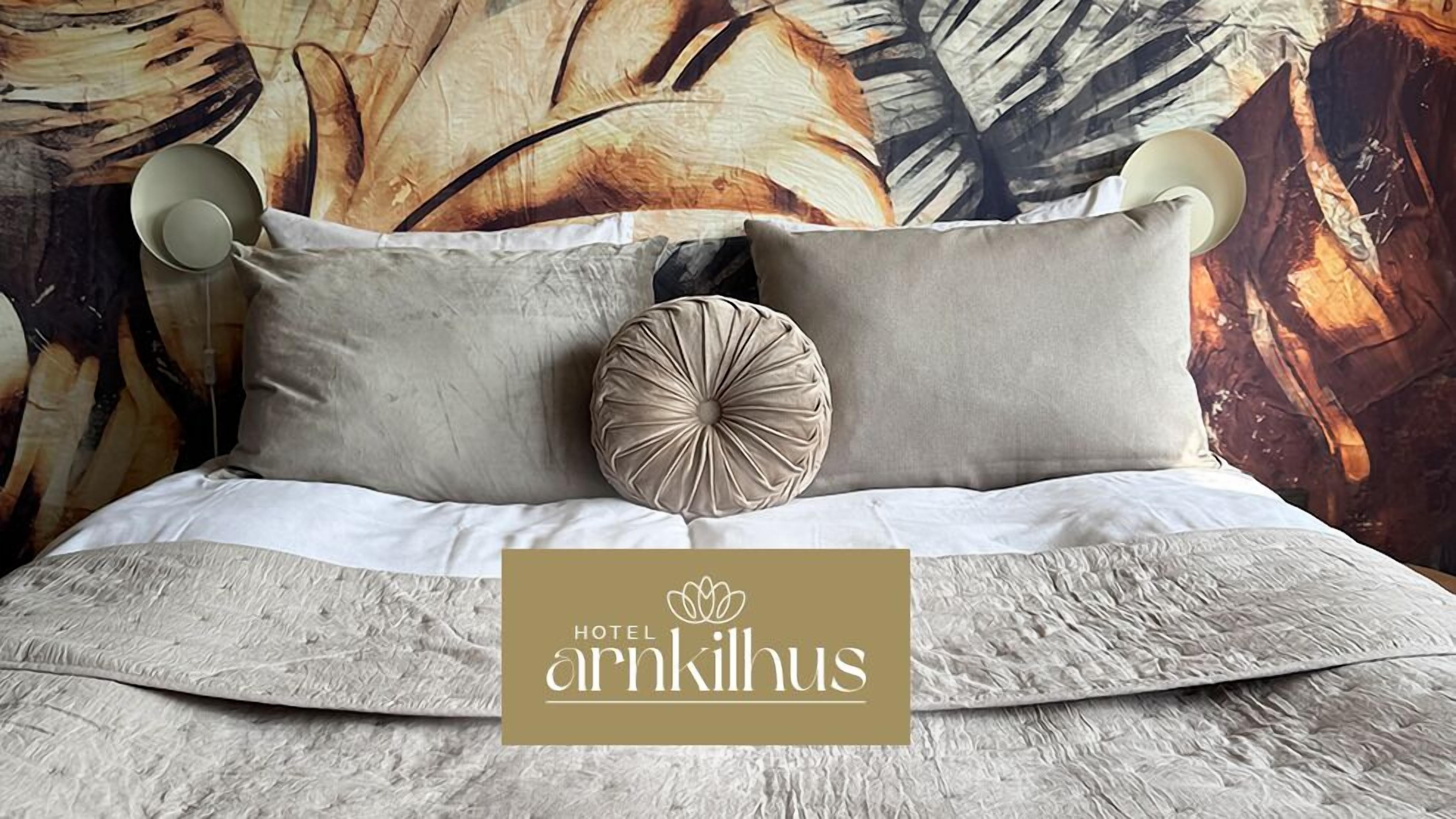 A bed with a textured white bedspread, two pillows, and a decorative round pillow. The background features a large abstract painting with leaf patterns.
