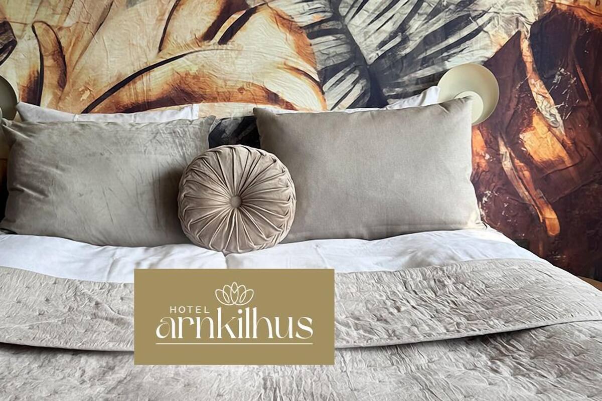 Un lit recouvert d ’une courtepointe blanche à motifs, avec deux oreillers et un coussin rond décoratif. Le fond est orné d’un grand tableau abstrait aux motifs de feuilles.