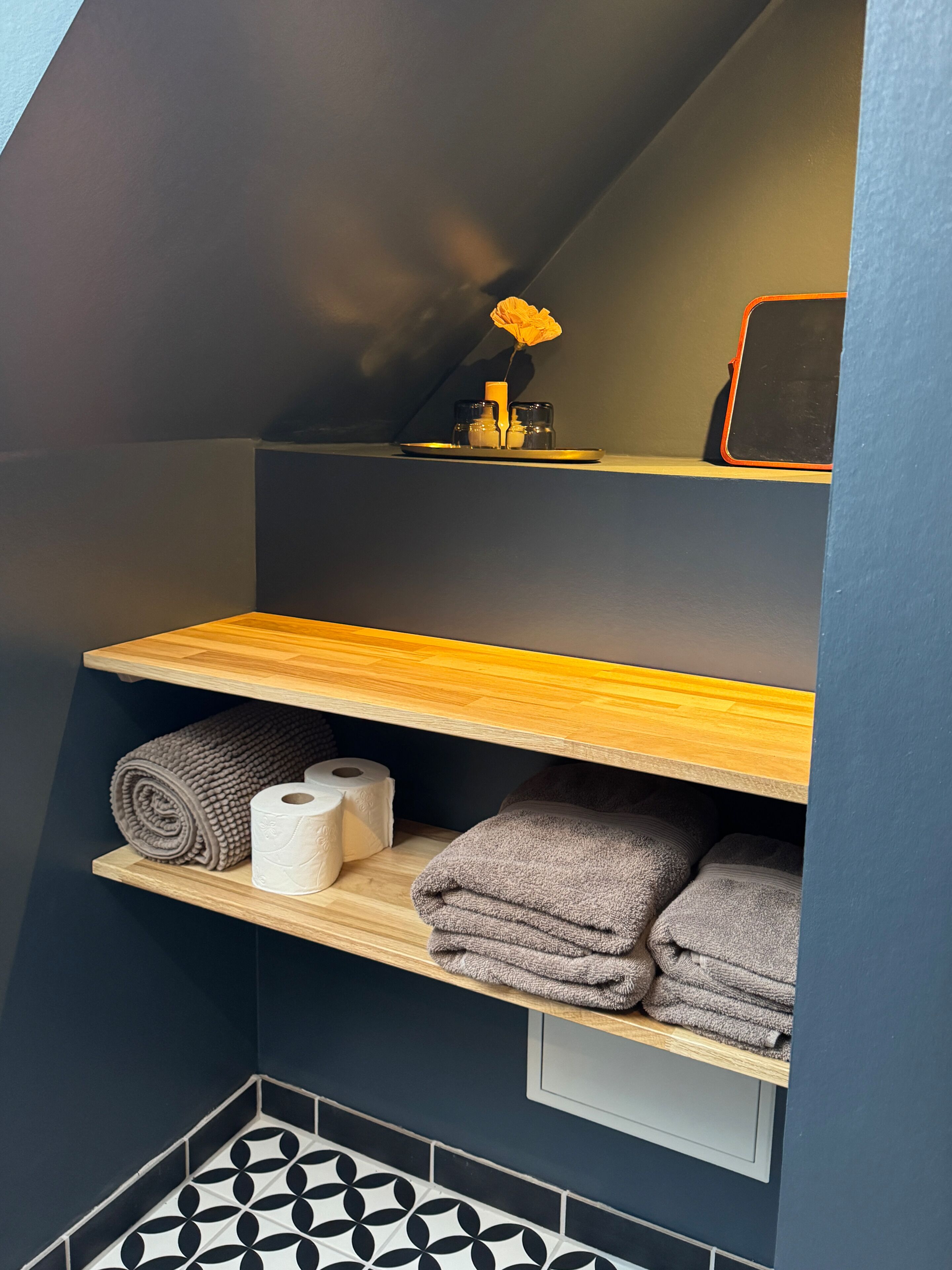 A bathroom with a wooden shelf, towels, toilet paper, and a small vase with a yellow flower.