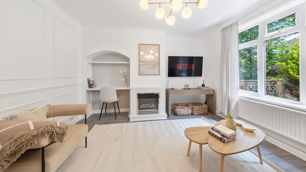 A living room with a beige sofa, a coffee table, a fireplace, a TV, and a large window with a view of greenery. - Modern 2-Bedroom Home in Bromley (Bromley)