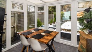 A conservatory with a wooden table and white chairs, large windows, and a view of a snowy garden. - Modern 2-Bedroom Home in Bromley (Bromley)