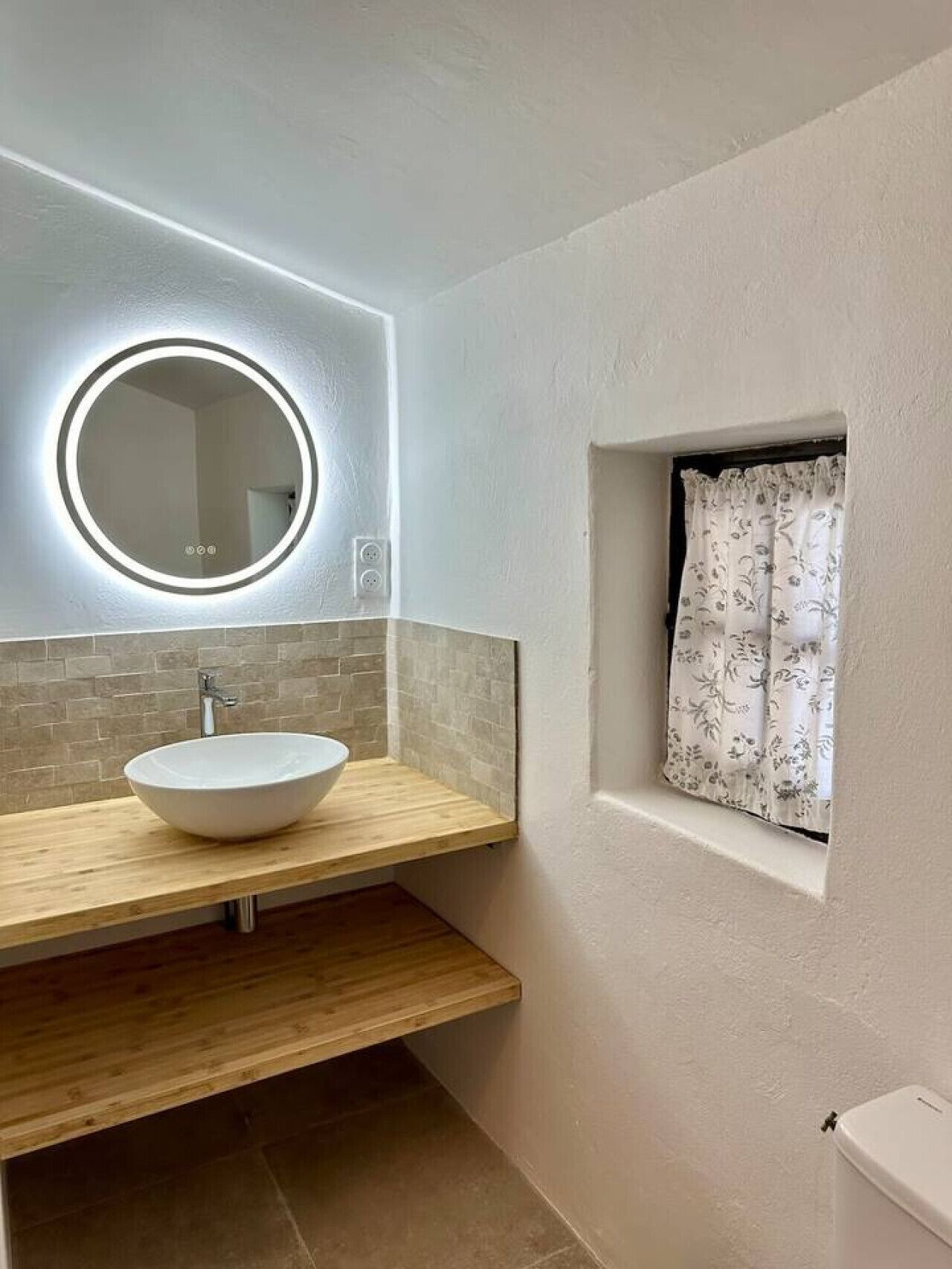 A bathroom with a round mirror, a white sink, and a window with a floral curtain.