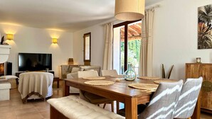 A dining area with a wooden table, cushioned chairs, and a television in the background. - Les Lauriers Blancs, air-conditioned villa with swimming pool (L'Isle-sur-la-Sorgue)