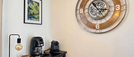 Une horloge murale au design circulaire, un tableau encadré, une machine à café, une bouilloire et une lampe, le tout posé sur une table noire.