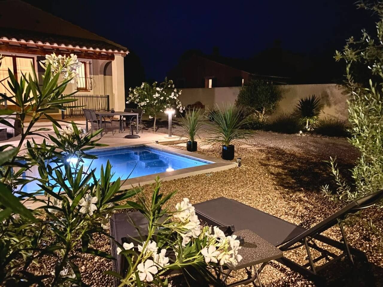 Une piscine entourée de chaises longues, de plantes et d’une maison en arrière-plan.