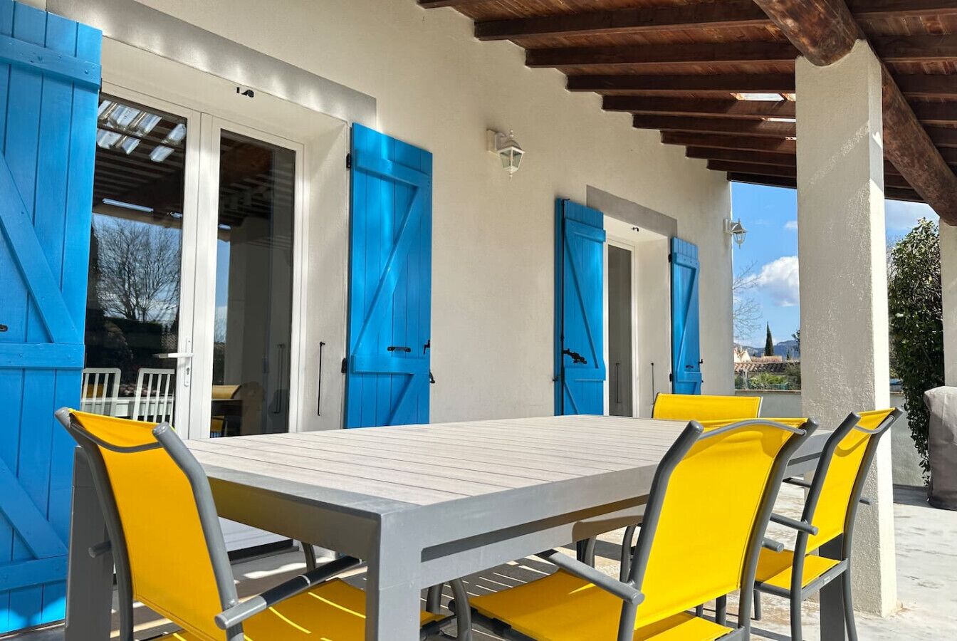 A covered outdoor dining area with a table and chairs, blue shutters, and a pergola structure.