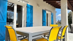 Ein überdachter Essbereich im Freien mit Tisch und Stühlen, blauen Fensterläden und einer Pergola.