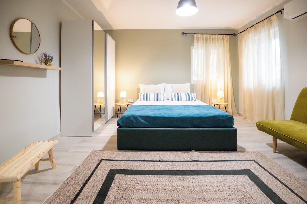 Une chambre moderne avec un grand lit, des lampes de chevet, un banc et un fauteuil vert.