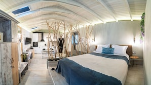 A modern bedroom with a large bed, a wooden bench, a small table, and a kitchen area in the background. - Vayas Residences Legacy (Trikala)