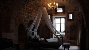 Una habitación tenuemente iluminada con una pared de piedra, una cama con dosel, una silla de madera y un banco.
