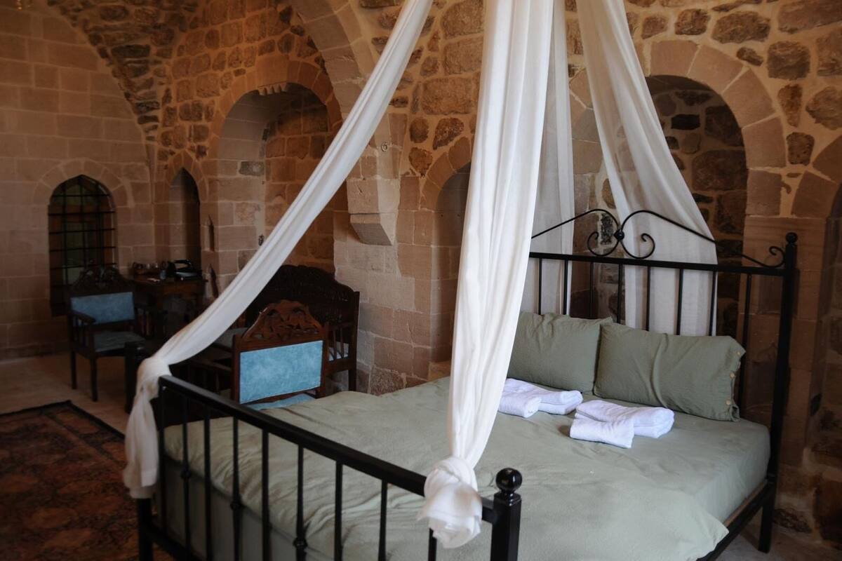 Une pièce aux murs de pierre, avec un lit à baldaquin, une chaise en bois et une petite table.