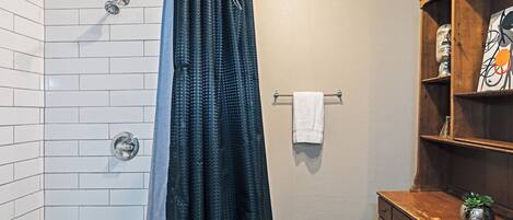 A bathroom with white tiled walls, a shower with a black shower curtain, a wooden cabinet with a plant, and a towel rack with a white towel.