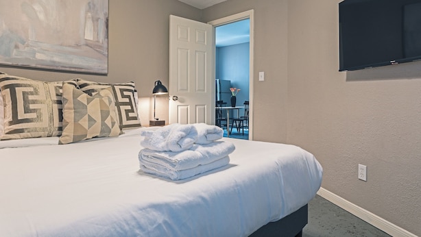 A neatly made bed with white linens and a stack of folded towels, a painting on the wall, a lamp, and a television mounted on the wall.