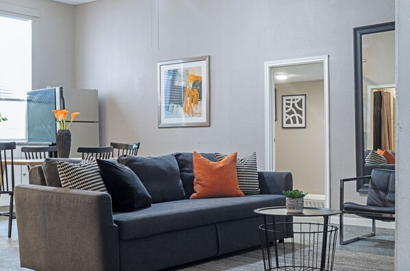 A modern living room with a grey sofa, a small round table, and a potted plant. - 3BR The Cardinal D, Montrose Downtown, MedCenter (Houston)