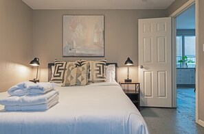 A neatly made bed with folded towels, a painting on the wall, and a door leading to another room. - 3BR The Cardinal D, Montrose Downtown, MedCenter (Houston)