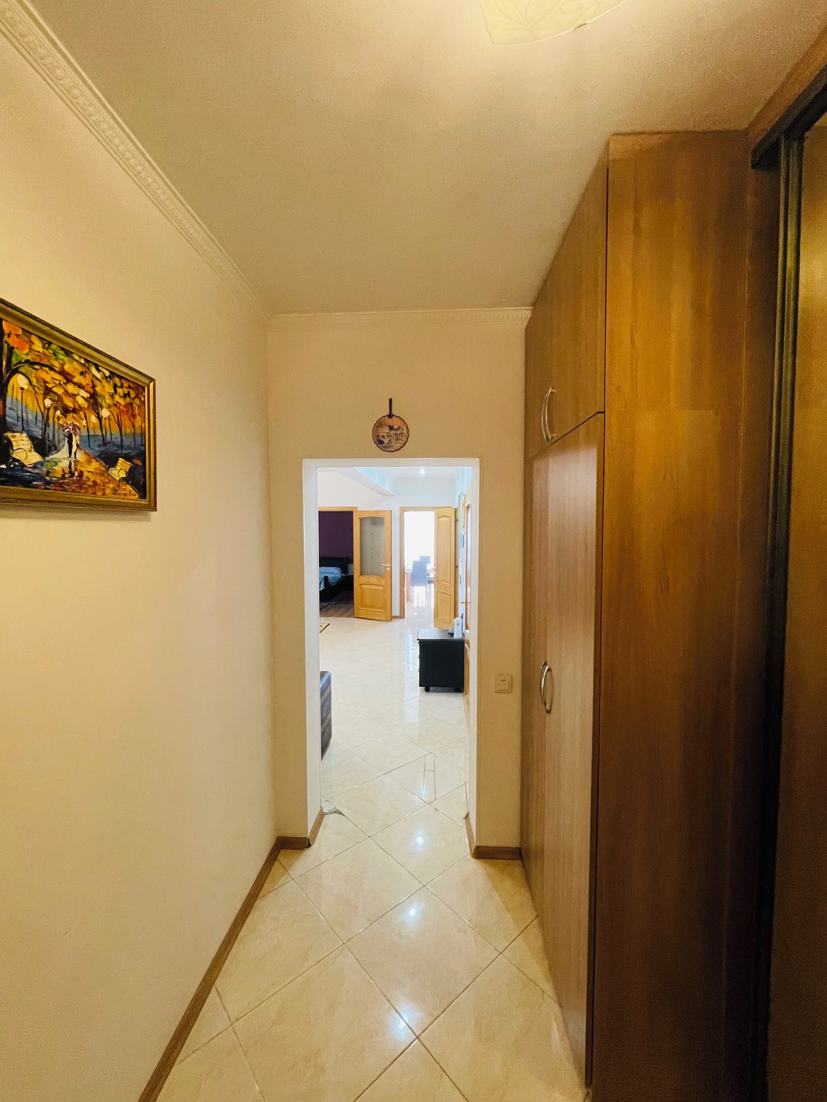 Un pasillo con puerta de madera, un cuadro en la pared y suelo de baldosas que conduce a una habitación con cama y escritorio.