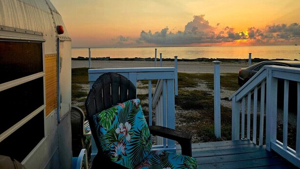 Une terrasse offrant une vue sur le coucher de soleil sur l’eau, une remorque abritée et une chaise avec un coussin à motifs floraux.