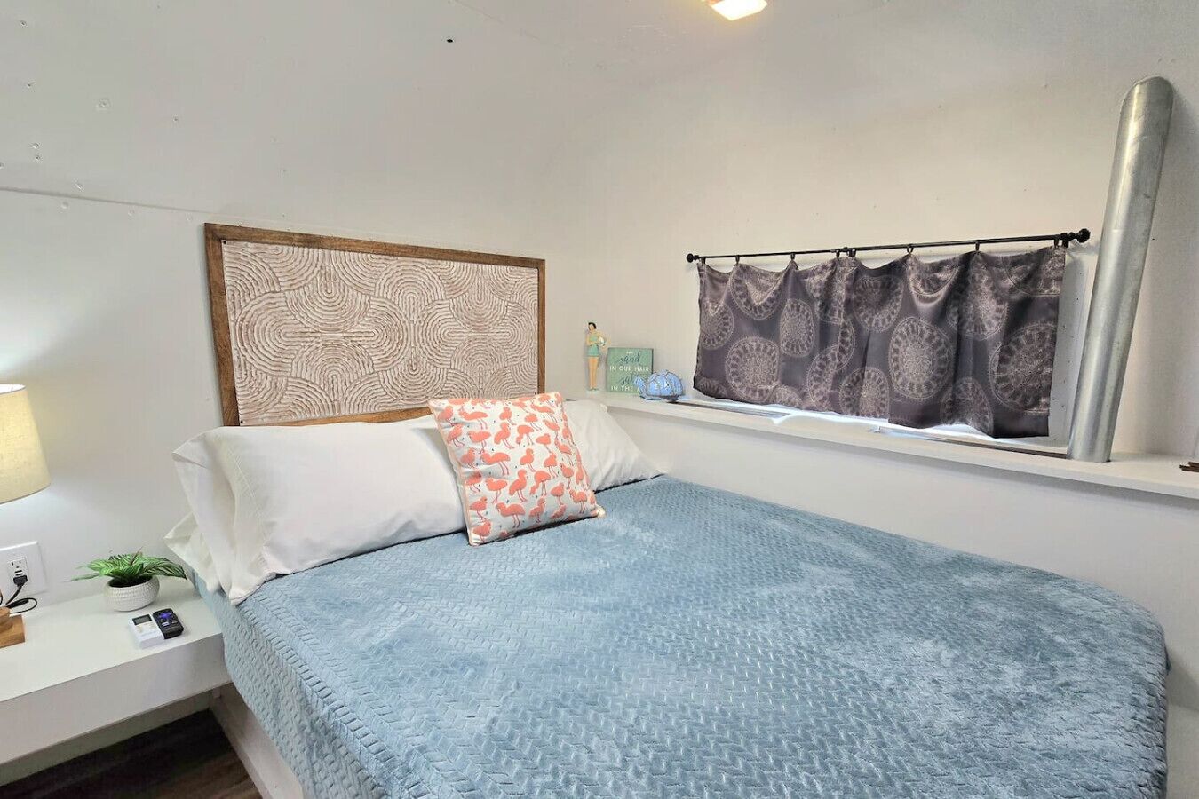 A neatly arranged bedroom with a bed, pillows, a bedside table with a lamp, a small plant, and a window with curtains.