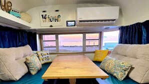 Un intérieur chaleureux avec une table en bois, deux canapés ornés de coussins à motifs, un climatiseur fixé au mur et une fenêtre offrant une vue sur le coucher de soleil.