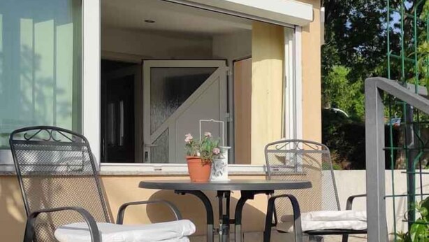 Eine Terrasse mit zwei Metallstühlen und einem Tisch, einem Blumentopf und einem Fenster mit Blick auf einen Garten.