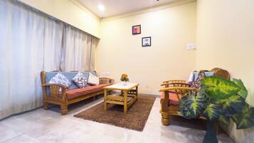 A living room with a sofa, two wooden chairs, a small table, a potted plant, and a ceiling fan.