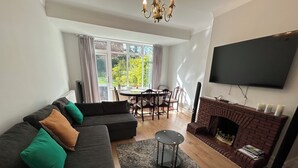 Een woonkamer met een bank, een open haard, een televisie en een eethoek met een tafel en stoelen.